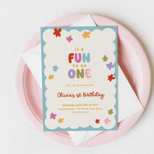 May include: A colorful invitation for Olivia's 1st birthday, featuring the text "It's FUN to be ONE" in playful, multi-colored letters. The invitation is decorated with flower illustrations and sits on a pink plate with a white envelope.