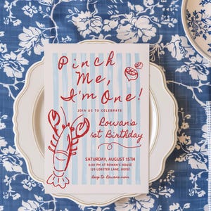 May include: A birthday invitation with the text "Pinch Me, I'm One!" and a red lobster illustration. The invitation is on a white plate, set on a blue floral tablecloth, with a knife and spoon.