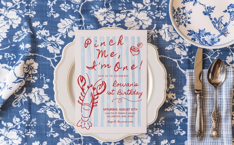 May include: A birthday invitation with the text "Pinch Me, I'm One!" and a red lobster illustration. The invitation is on a white plate, set on a blue floral tablecloth, with a knife and spoon.