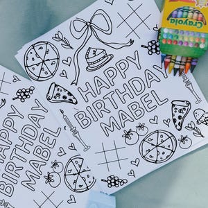 May include: Two white coloring pages with the text "HAPPY BIRTHDAY MABEL" and various line drawings of pizza, cake, and bows. A box of Crayola crayons is visible in the corner.
