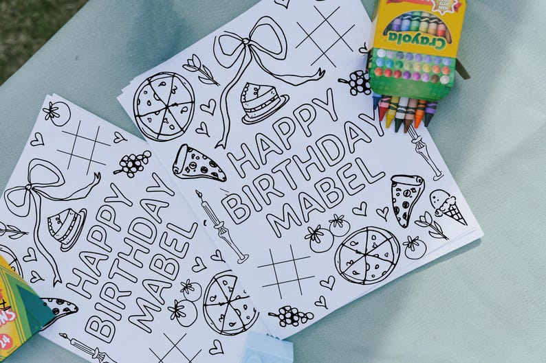 May include: Two white coloring pages with the text "HAPPY BIRTHDAY MABEL" and various line drawings of pizza, cake, and bows. A box of Crayola crayons is visible in the corner.