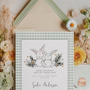 May include: A baby shower invitation with a green and white gingham background. Two white bunnies are surrounded by green leaves and yellow flowers. The text reads "Some bunnies are on their way. Join us for a baby shower honoring Sadie Anderson."