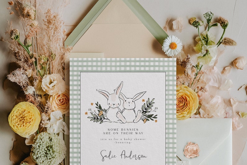 May include: A baby shower invitation with a green and white gingham background. Two white bunnies are surrounded by green leaves and yellow flowers. The text reads "Some bunnies are on their way. Join us for a baby shower honoring Sadie Anderson."