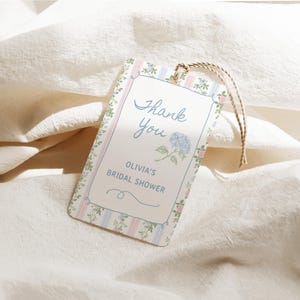 May include: A rectangular tag with a floral and striped border, featuring the text "Thank You" in cursive, and "Olivia's Bridal Shower." A blue hydrangea illustration is also present. The tag is attached to a natural twine string.