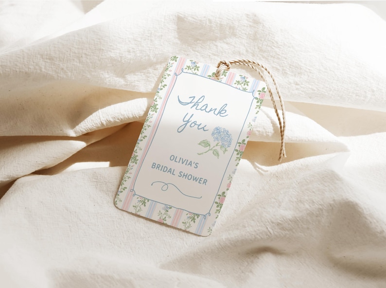 May include: A rectangular tag with a floral and striped border, featuring the text "Thank You" in cursive, and "Olivia's Bridal Shower." A blue hydrangea illustration is also present. The tag is attached to a natural twine string.