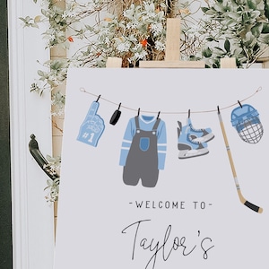 Pode incluir: Um cartaz de boas-vindas branco com ilustra&ccedil;&otilde;es de h&oacute;quei azul e cinza penduradas em um varal. O cartaz diz "Bem-vindo ao Taylor's".