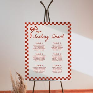 Pode incluir: Um diagrama de lugares a xadrez vermelho e branco com um laço vermelho no topo. O diagrama lista os nomes dos convidados para cada mesa, com cada número de mesa em vermelho.