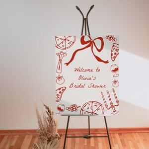May include: A white welcome sign with red handwritten text and illustrations of pizza, wine glasses, and grapes. The text reads "Welcome to Olivia's Bridal Shower". The sign is on an easel.