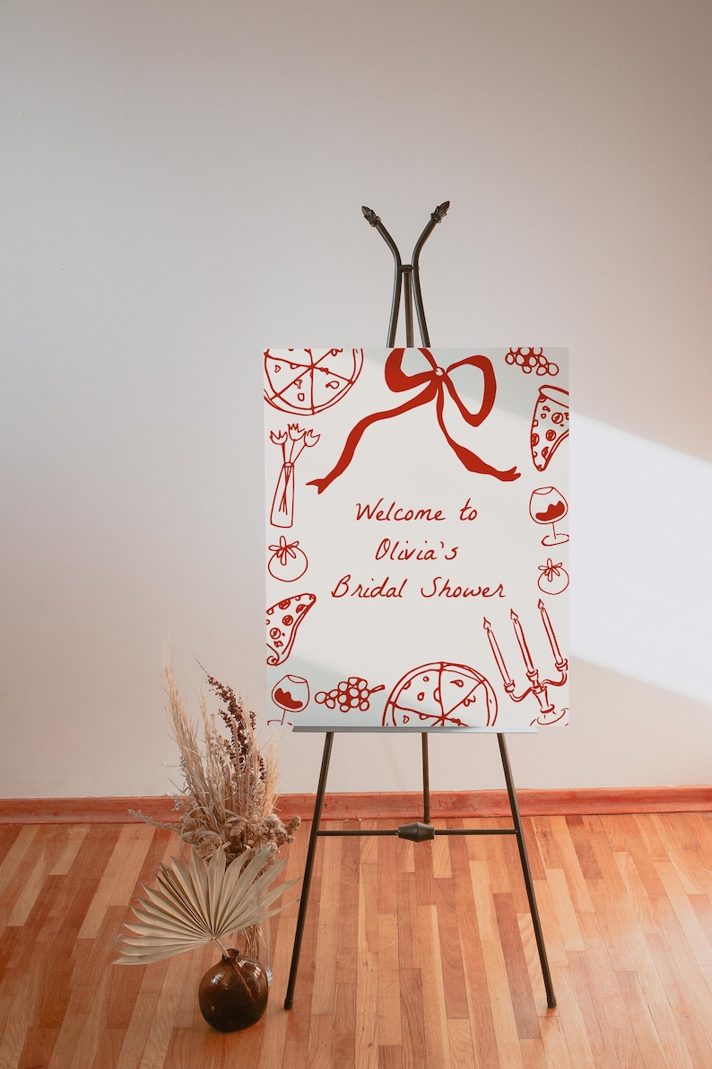 May include: A white welcome sign with red handwritten text and illustrations of pizza, wine glasses, and grapes. The text reads "Welcome to Olivia's Bridal Shower". The sign is on an easel.