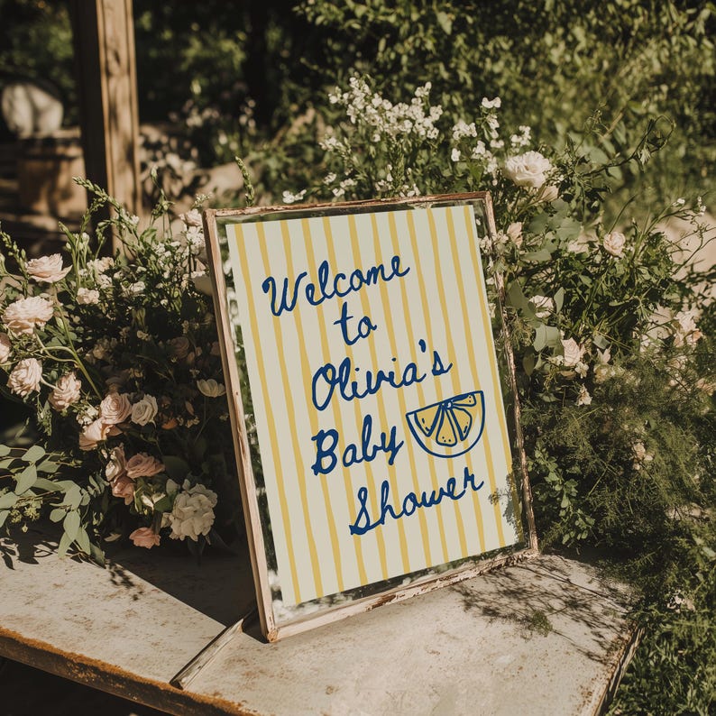 May include: A framed sign with the text "Welcome to Olivia's Baby Shower" in blue script. The sign has a yellow and white striped background and a lemon slice graphic. The sign is surrounded by greenery and flowers.