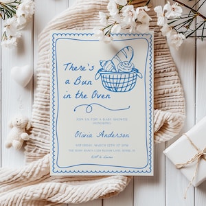 May include: A blue and white baby shower invitation with the text "There's a Bun in the Oven" and a blue and white illustration of a basket of bread. The invitation includes the date, time, and location of the shower.
