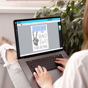 May include: A person using a laptop to design a digital file. The design features a skeleton hand making a peace sign with the text "Oscar is bad two the bone".