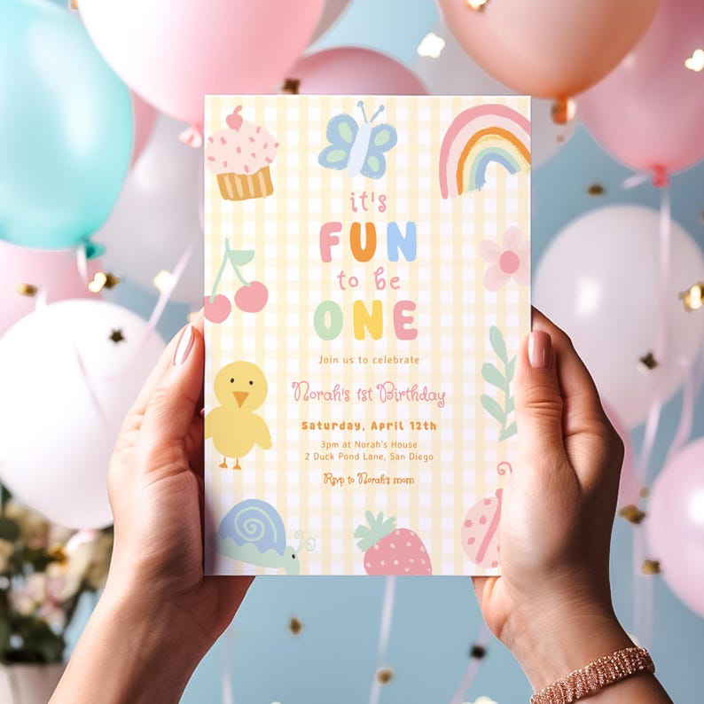 May include: A birthday invitation held by hands, featuring the text "it's FUN to be ONE." The invitation has a yellow gingham background with colorful illustrations of a cupcake, butterfly, rainbow, and other playful elements. The invitation is for Norah's 1st birthday.