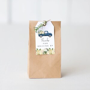 May include: A brown paper bag with a white tag attached. The tag has a blue pickup truck with flowers in the back. The text on the tag reads "Thanks for driving by".