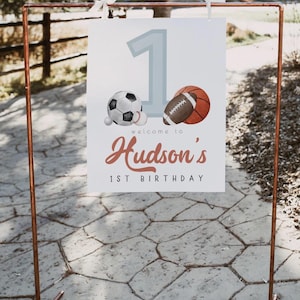 May include: A white sign with a blue number one, a soccer ball, a baseball, and a basketball. The sign says "Welcome to Hudson's 1st Birthday".