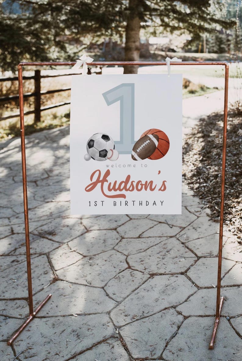 May include: A white sign with a blue number one, a soccer ball, a baseball, and a basketball. The sign says "Welcome to Hudson's 1st Birthday".