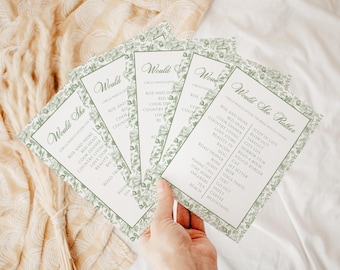 Op de afbeelding: Een hand houdt een waaier van vijf witte kaarten met groene bloemenranden vast. Elke kaart bevat de tekst "Would She Rather" en een lijst met opties. De kaarten zijn gerangschikt op een getextureerd, lichtgekleurd oppervlak.
