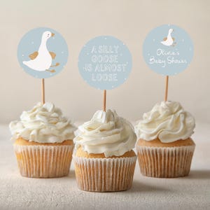 May include: Three cupcakes with white frosting and blue and white circular toppers. The toppers feature a cartoon goose, the text "A silly goose is almost loose", and the text "Olivia's Baby Shower".