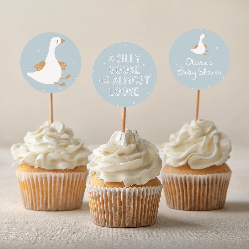 May include: Three cupcakes with white frosting and blue and white circular toppers. The toppers feature a cartoon goose, the text "A silly goose is almost loose", and the text "Olivia's Baby Shower".