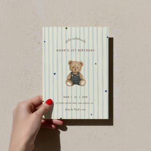 May include: An invitation card with a light blue and white striped design, featuring a teddy bear illustration and the text "Let's celebrate" and "Noah's 1st Birthday". The card includes event details and is held by a hand.