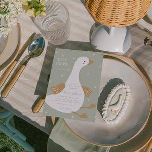 Op de afbeelding: Een baby shower uitnodiging met een cartoon gans op een groene achtergrond. De uitnodiging zegt "A Silly Goose" en bevat de naam "Olivia Johnson" en de datum "Saturday, April 15th".