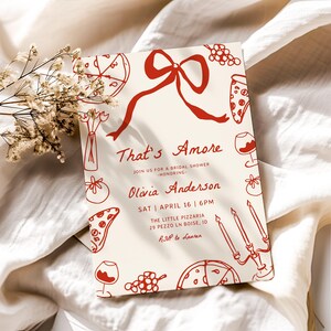 May include: A red and white bridal shower invitation with the text "That's Amore" and "Olivia Anderson". The invitation features a red bow, pizza slices, wine glasses, grapes, and candles.
