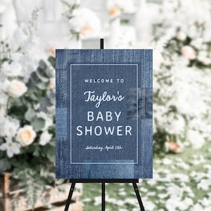 May include: A denim-textured sign on a black easel, announcing a baby shower. The sign reads "Welcome to Taylor's BABY SHOWER" with the date "Saturday, April 15th" below. The background is blurred with floral arrangements.