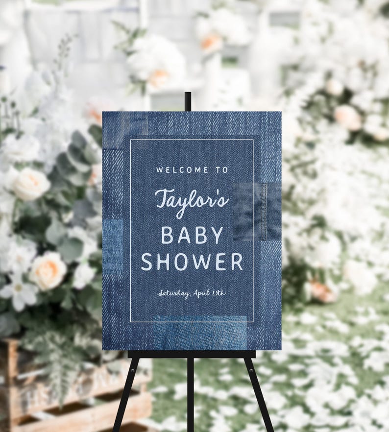 May include: A denim-textured sign on a black easel, announcing a baby shower. The sign reads "Welcome to Taylor's BABY SHOWER" with the date "Saturday, April 15th" below. The background is blurred with floral arrangements.
