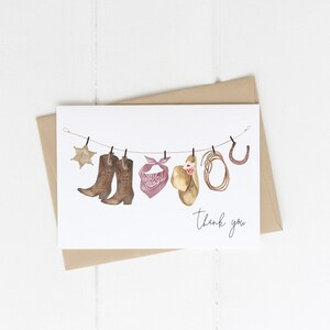 May include: A white card with a thank you message and a string of cowboy-themed items hanging from it. The items include a sheriff's star, two brown cowboy boots, a pink bandana, a brown cowboy hat, a brown rope, and a horseshoe.