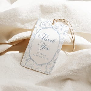 Op de afbeelding: Een rechthoekig kraamcadeau-label met een lichtblauw bloemmotief en de woorden "Thank You" in elegant schrift. Het label bevat de tekst "Olivia's Baby Shower" en is vastgebonden met een natuurlijk touw.