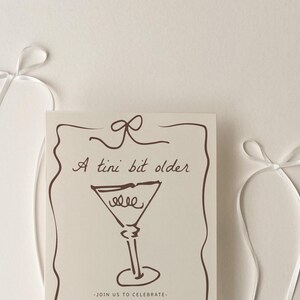 Op de afbeelding: Een witte kaart met bruine tekst en een tekening van een martini-glas. De tekst luidt "A tini bit older" en "Join us to celebrate".