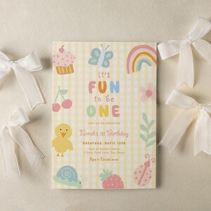 May include: A birthday invitation with the text "it's FUN to be ONE" on a yellow gingham background. The invitation is decorated with colorful illustrations of a cupcake, butterfly, rainbow, and other cute graphics. White satin bows are scattered around the invitation.