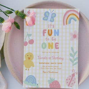 May include: A pastel-colored birthday invitation with the text "it's FUN to be ONE." The invitation features illustrations of a butterfly, rainbow, cherries, chick, snail, strawberry, and ladybug. The invitation is on a pink plate with pink flowers.
