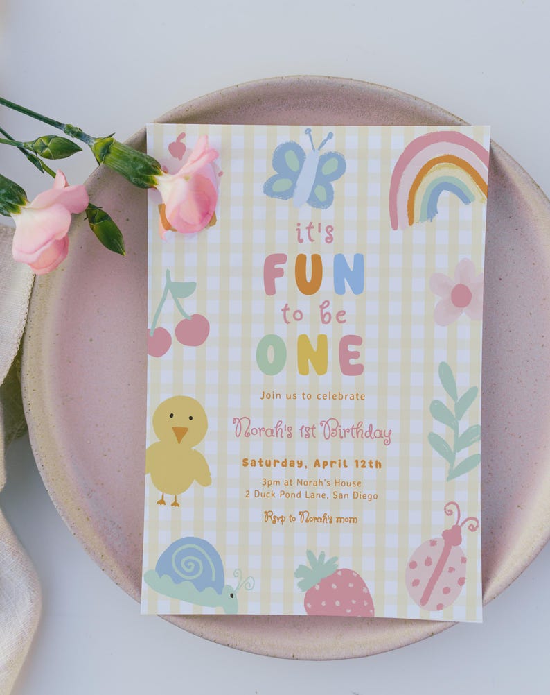 May include: A pastel-colored birthday invitation with the text "it's FUN to be ONE." The invitation features illustrations of a butterfly, rainbow, cherries, chick, snail, strawberry, and ladybug. The invitation is on a pink plate with pink flowers.