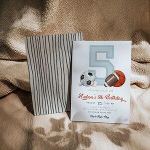 May include: A birthday invitation with a light blue number 5 and sports ball illustrations. The invitation includes the text "Let's have a ball for Hudson's 5th Birthday" and event details. A striped paper bag is next to the invitation.