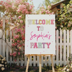 Peut inclure: Panneau d'accueil pour la fête de Sophia avec des lettres scintillantes colorées sur fond blanc. Le panneau est sur un chevalet devant une clôture blanche et des fleurs roses.