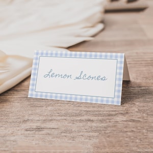 May include: A folded place card with a blue gingham border and the words "Lemon Scones" in cursive script. The card sits on a wooden surface, with a white, folded paper element in the background.