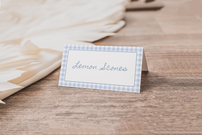 May include: A folded place card with a blue gingham border and the words "Lemon Scones" in cursive script. The card sits on a wooden surface, with a white, folded paper element in the background.