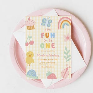 May include: A pastel-colored birthday invitation with the text "It's FUN to be ONE" on a yellow gingham background. The invitation includes illustrations of a cupcake, butterfly, rainbow, and other cute graphics. It rests on a pink plate.