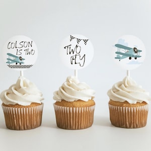 May include: Three cupcakes with white frosting and round toppers. The toppers feature a blue airplane, the text "Colson is Two" and the text "Two Fly".