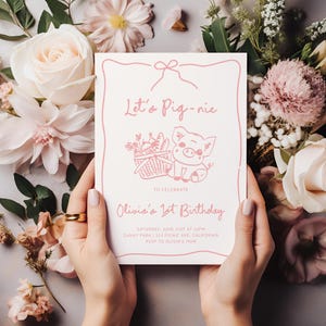 May include: A pink invitation with the text "Let's Pig-nic" and "Olivia's 1st Birthday". The invitation features a cartoon pig with a picnic basket. The invitation is held by hands, surrounded by pink and white flowers.