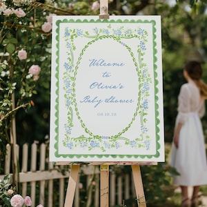 Puede incluir: Un letrero en un caballete con el texto "Welcome to Olivia's Baby Shower" en escritura azul. El letrero tiene un borde verde festoneado y un diseño floral ovalado. La fecha "08.10.28" está en la parte inferior. El fondo muestra un jardín.