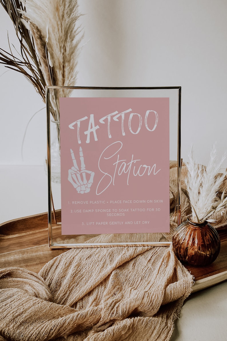 Puede incluir: Un cartel con marco dorado, fondo rosa y texto blanco que dice "TATTOO Station". El cartel incluye instrucciones para aplicar tatuajes temporales: 1. Quitar el pl&aacute;stico - Colocar la cara hacia abajo sobre la piel. 2. Usar una esponja h&uacute;meda para remojar el tatuaje durante 30 segundos. 3. Levantar el papel suavemente y dejar secar.