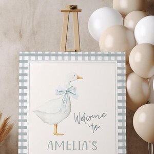 May include: A welcome sign with a gray and white checkered border. The sign features a watercolor illustration of a white goose with a blue bow. The text on the sign reads "Welcome to Amelia's".