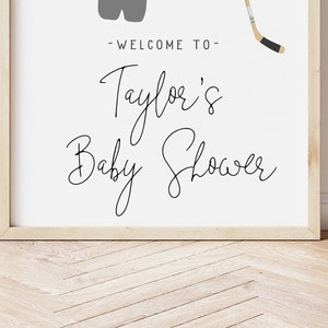 Pode incluir: Uma placa branca emoldurada com texto preto que diz "Bem-vindo ao Chuveiro de Beb&ecirc; de Taylor". Um taco de h&oacute;quei e um par de patins de gelo s&atilde;o ilustrados em cinza na placa.