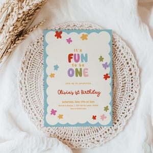 May include: A light blue birthday invitation with a scalloped edge, featuring the text "it's FUN to be ONE" in colorful letters. The invitation includes details for Olivia's 1st birthday, surrounded by flower illustrations.