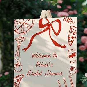 May include: A white sign with red illustrations of pizza, a bow, a wine glass, and other food items. The sign reads "Welcome to Olivia's Bridal Shower".