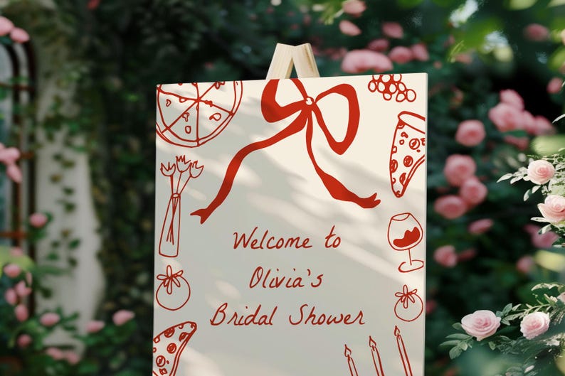 May include: A white sign with red illustrations of pizza, a bow, a wine glass, and other food items. The sign reads "Welcome to Olivia's Bridal Shower".