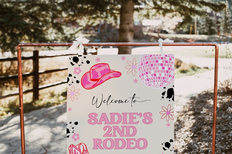 May include: A pink and white welcome sign with the text "Welcome to Sadie's 2nd Rodeo". The sign has a pink cowboy hat, a pink disco ball, and black and white cow print accents.