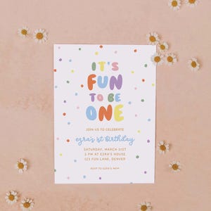 May include: A white invitation card with the text "IT'S FUN TO BE ONE" in colorful letters and confetti dots. The card announces Ezra's 1st birthday party, including the date, time, and location. Small white daisies are scattered around the card.